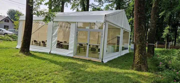 Mały biały pawilon namiotowy z przeszklonymi drzwiami, stołami i krzesłami, rozstawiony na trawie między drzewami
