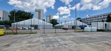Dwie białe hale namiotowe na utwardzonym placu między wysokimi blokami